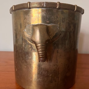 Puede incluir: Un cubo de hielo de metal dorado con un diseño de cabeza de elefante en la parte delantera. El cubo tiene un borde decorativo y está hecho de un metal brillante.