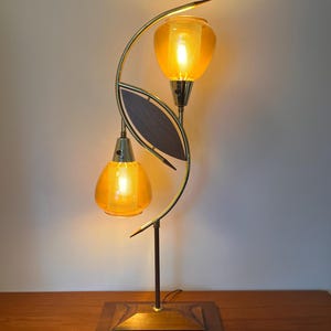 Könnte beinhalten: Eine moderne Tischlampe aus der Mitte des Jahrhunderts mit einem Sockel aus Messing und Holz und zwei Schirmen aus bernsteinfarbenem Glas. Die Lampe hat ein einzigartiges geschwungenes Design und wird mit warmweißem Licht beleuchtet.