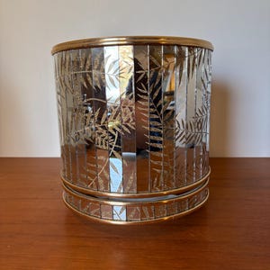 Może przedstawiać: Cylindryczny pojemnik dekoracyjny z lustrzaną powierzchnią. Pojemnik posiada złoty rant i podstawę, z powtarzającym się wzorem złotych liści. Pojemnik ma około 25 cm wysokości i 20 cm średnicy.
