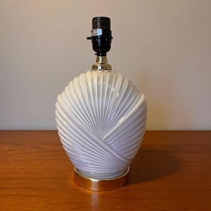 Op de afbeelding: Witte keramische tafellamp met een gouden voet en een waaiervormig ontwerp. De lamp heeft een wit snoer en een zwarte fitting.