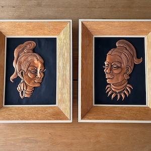 Puede incluir: Dos esculturas en relieve de cobre enmarcadas de rostros de mujeres. La obra de arte de cobre está sobre un fondo negro y enmarcada en madera clara. Una mujer lleva pendientes, la otra un collar. Los marcos tienen un aspecto texturizado.