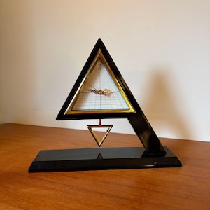 May include: A black and gold geometric desk clock. The clock face is square with gold hands and Roman numerals. The clock is in a triangular frame with a smaller triangle suspended below.