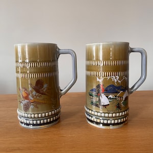 Puede incluir: Dos tazas de cerámica verde con asas. Una taza presenta un diseño de faisán y la otra una mujer caminando por un campo con una cabaña en el fondo. La taza con la mujer tiene la palabra "Irlanda" impresa en la parte inferior.