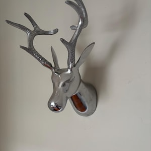 Puede incluir: Una cabeza de ciervo plateada para colgar en la pared, con cuernos detallados y un acabado pulido. La cabeza de ciervo está montada en una pared blanca, creando un acento decorativo llamativo. Una escultura moderna de un trofeo de caza clásico.