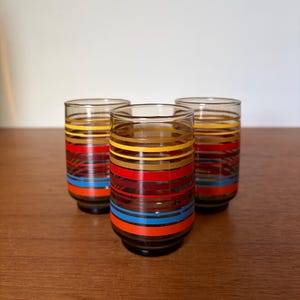 May include: Three cylindrical drinking glasses made of translucent brown glass. Each glass features a series of horizontal stripes in vibrant colours including yellow, orange, red, and bright blue, wrapping around the exterior surface.
