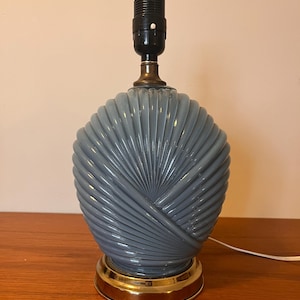 以下が含まれることがあります： 扇形のテクスチャードボディと真鍮色のベースを備えたグレーのテーブルランプ。 ランプには黒いソケットと白いコードが付いています。 ランプは木製の表面にあります。