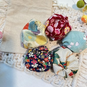 May include: A set of five small, round fabric pouches featuring various floral patterns in red, blue, green, and gold, accompanied by a plain beige drawstring bag. The pouches are crafted from patterned textile with intricate chrysanthemum and cherry blossom motifs.