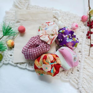 May include: A collection of five small, round fabric pouches featuring various Japanese-inspired floral and geometric patterns in red, purple, pink, and white. These soft, textured textile items are arranged alongside a plain beige drawstring bag.