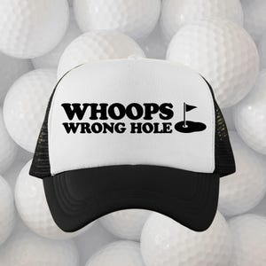 May include: A black and white trucker hat with the text "WHOOPS WRONG HOLE" in bold, black letters. A small graphic of a golf hole with a flag is to the right of the text. The hat has a black brim and mesh back.