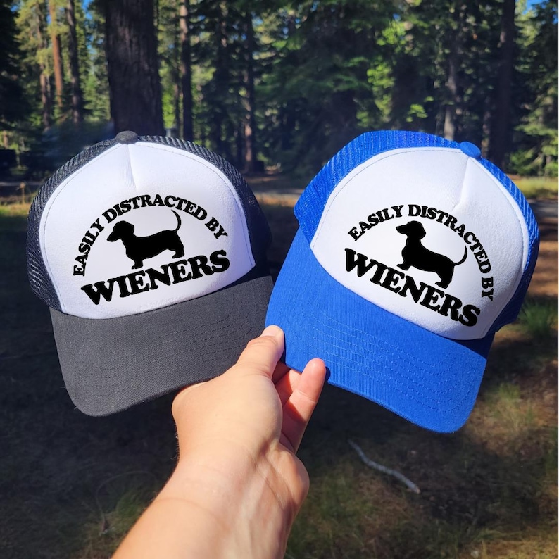 K&ouml;nnte beinhalten: Zwei schwarze und wei&szlig;e Trucker-Caps mit einer blauen und wei&szlig;en Trucker-Cap. Die Caps haben einen wei&szlig;en Hintergrund mit schwarzem Text, der "Easily Distracted by Wieners" lautet, und eine schwarze Silhouette eines Dackels.