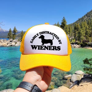 K&ouml;nnte beinhalten: Eine gelbe und wei&szlig;e Trucker-Kappe mit einer schwarzen Dackel-Silhouette und dem Text "Easily Distracted by Wieners".