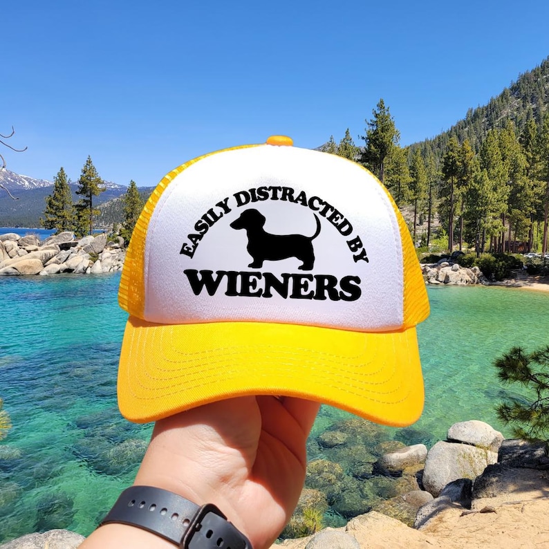 K&ouml;nnte beinhalten: Eine gelbe und wei&szlig;e Trucker-Kappe mit einer schwarzen Dackel-Silhouette und dem Text "Easily Distracted by Wieners".