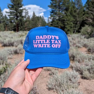 May include: A bright blue trucker cap featuring a mesh back and a solid front panel. The text DADDY'S LITTLE TAX WRITE OFF is printed in bold pink capital letters across the centre of the front panel. The cap has a curved brim and a matching blue cord.