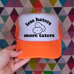 May include: A bright orange trucker hat with a white front panel featuring a black graphic of two potatoes and the text "less haters more taters" printed in a bold, rounded font. The cap includes a mesh back and a solid orange brim.