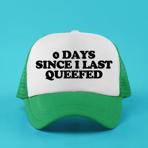 May include: A green and white trucker hat with the text "0 DAYS SINCE I LAST QUEEFED" in black. The hat has a white front panel, a green brim, and a green mesh back. The background is a solid light blue.