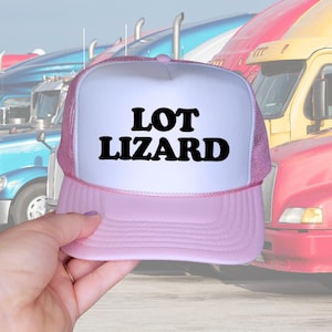 May include: A light pink and white trucker hat featuring a mesh back and a solid front panel. The text "LOT LIZARD" is printed in bold, black, rounded lettering across the center of the white front panel of the cap.