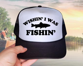 Puede incluir: Una gorra tipo trucker negra y blanca con un panel frontal blanco que presenta el texto negro "WISHIN' I WAS FISHIN'" centrado sobre y debajo de una silueta negra de un pez. La gorra tiene una visera negra y paneles laterales de malla.