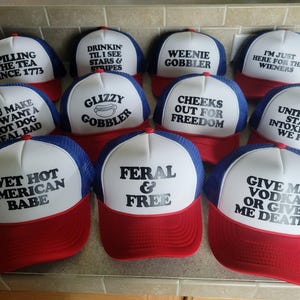 K&ouml;nnte beinhalten: Eine Sammlung von zehn rot-wei&szlig;-blauen Trucker-Caps mit verschiedenen schwarzen Text-Slogans, darunter Spilling The Tea Since 1773, Drinkin Til I See Stars & Stripes, Weenie Gobbler, I'm Just Here For The Wieners und Feral & Free.