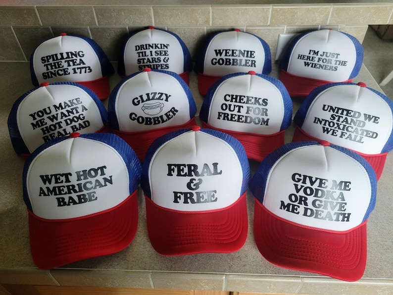 K&ouml;nnte beinhalten: Eine Sammlung von zehn rot-wei&szlig;-blauen Trucker-Caps mit verschiedenen schwarzen Text-Slogans, darunter Spilling The Tea Since 1773, Drinkin Til I See Stars & Stripes, Weenie Gobbler, I'm Just Here For The Wieners und Feral & Free.