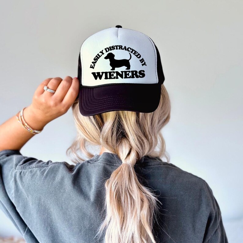 K&ouml;nnte beinhalten: Schwarz-wei&szlig;e Trucker-Kappe mit wei&szlig;em Netzr&uuml;cken. Die Kappe hat eine schwarze Dackel-Silhouette und den Text "Easily Distracted by Wieners".