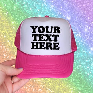 May include: A bright pink and white trucker cap featuring a foam front panel with the black text YOUR TEXT HERE printed in a bold, rounded font. The cap includes a matching pink mesh back and a solid pink brim with horizontal stitching details.