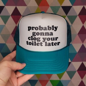 May include: A teal and white trucker hat with the text "probably gonna clog your toilet later" in black. The hat has a white front panel, a teal brim, and a teal mesh back. The text is centered on the front panel.