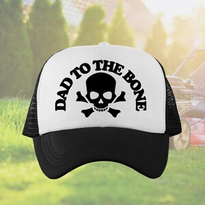 May include: A black and white trucker hat featuring the text "DAD TO THE BONE" above a skull and crossbones. The hat has a white front panel, a black peak, and a black mesh back.