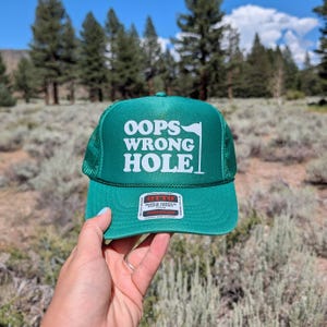 May include: A green trucker hat with a mesh back and a foam front panel. The front features white text that reads OOPS WRONG HOLE with a small golf flag icon. A rectangular sticker on the brim displays the brand name OTTO and product details.