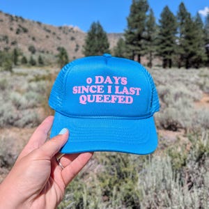 May include: A bright blue trucker hat featuring a foam front panel and a mesh back. The front of the hat displays the text 0 DAYS SINCE I LAST QUEEFED printed in bold, light pink, sans-serif lettering. The hat includes a matching blue brim.