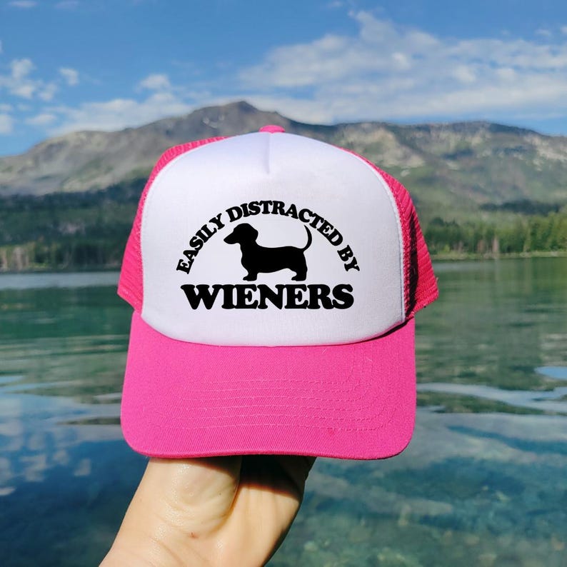 K&ouml;nnte beinhalten: Eine rosafarbene und wei&szlig;e Trucker-Kappe mit einer schwarzen Silhouette eines Dackels und dem Text "Easily Distracted by Wieners".