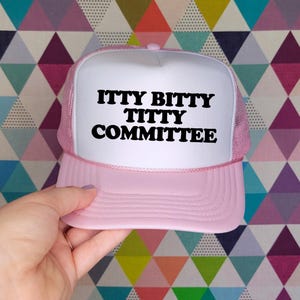May include: A light pink and white trucker cap featuring a foam front panel with the black text ITTY BITTY TITTY COMMITTEE printed in a bold, rounded font. The cap includes a matching pink mesh back, a curved peak, and a pink cord trim detail.