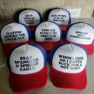 K&ouml;nnte beinhalten: Eine Sammlung von sieben rot-wei&szlig;-blauen Trucker-Caps mit Schaumstofffront und Netzr&uuml;ckseite. Jede Kappe zeigt einen einzigartigen schwarzen Text auf der wei&szlig;en Vorderseite, darunter Phrasen wie Star Spangled & Spread Eagle und Bitches Love Independence.