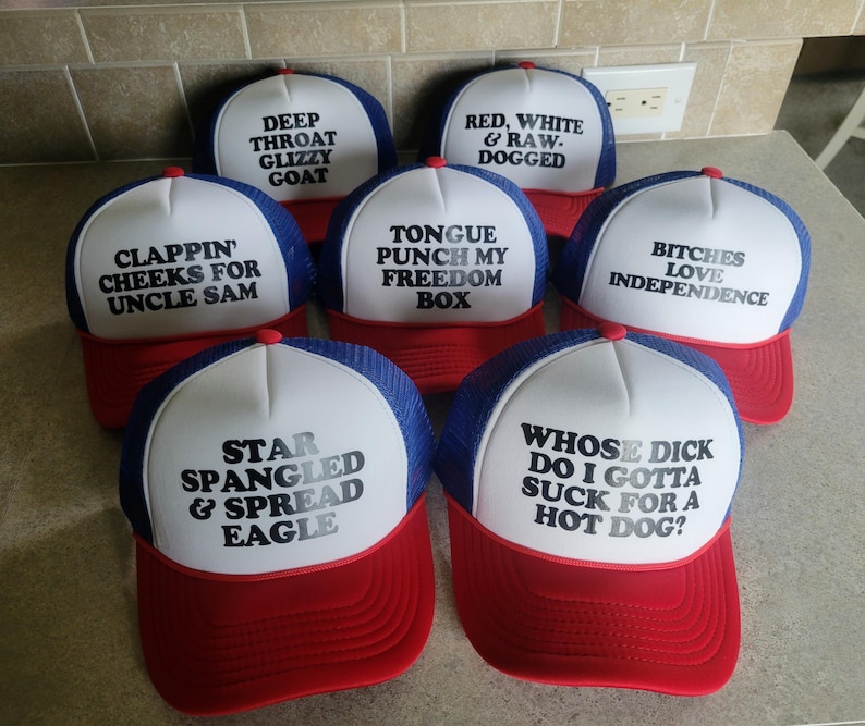 K&ouml;nnte beinhalten: Eine Sammlung von sieben rot-wei&szlig;-blauen Trucker-Caps mit Schaumstofffront und Netzr&uuml;ckseite. Jede Kappe zeigt einen einzigartigen schwarzen Text auf der wei&szlig;en Vorderseite, darunter Phrasen wie Star Spangled & Spread Eagle und Bitches Love Independence.