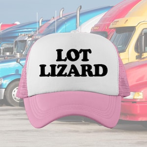 May include: A trucker hat with a white front panel and a pink brim and mesh back. The text "LOT LIZARD" is printed in bold black letters on the front panel. The background features a row of semi-trucks.