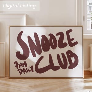 May include: A framed art print with the words "SNOOZE CLUB" in a bold, dark red font. Below, the text "7 AM DAILY" is also in dark red. The artwork is displayed on a light beige background, with a wooden frame.