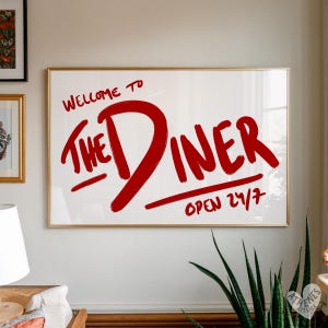 May include: A framed print with the words "WELCOME TO THE DINER" in red brushstrokes on a white background. Below the diner text is the phrase "OPEN 24/7". The frame is gold.