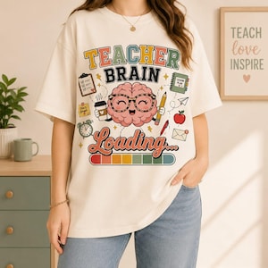 May include: An off-white t-shirt featuring a graphic of a cartoon brain wearing glasses, holding a coffee cup and pencil. The text reads TEACHER BRAIN Loading... surrounded by icons of a clipboard, lesson plan, apple, and a loading progress bar.