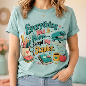 May include: A light blue t-shirt featuring a graphic print with the text Everything Has A Home... Except My Stapler. The design includes a pencil holder with stationery, a spiral notebook, a coffee mug, an apple, and a cartoon stapler with legs.