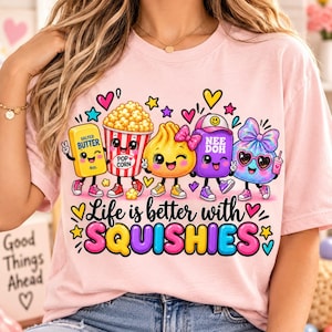 Puede incluir: Una camiseta rosa claro con un gráfico de cinco personajes coloridos y antropomórficos, incluyendo una barra de mantequilla, palomitas, una empanadilla, un bloque morado con la etiqueta NEE DOH y un objeto con lazo azul, con el texto Life is better with SQUISHIES.