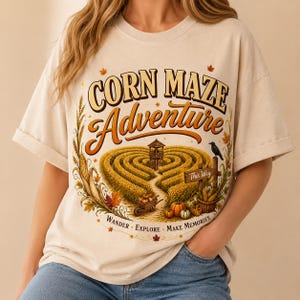 Op de afbeelding: Een crèmekleurig t-shirt met een afbeelding van een maïsdoolhof, een uitkijktoren, pompoenen en een zwarte kraai. De tekst luidt "CORN MAZE Adventure" en "WANDER EXPLORE MAKE MEMORIES" met "This Way" op een houten bord, omringd door herfstbladeren.
