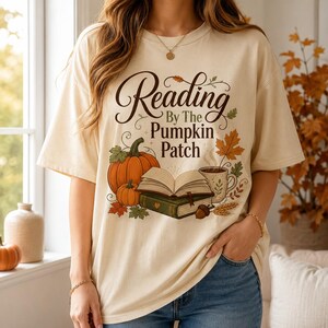 May include: A cream-colored short-sleeve t-shirt featuring a graphic print with the text Reading By The Pumpkin Patch, an open book resting on a closed book, a large orange pumpkin, a smaller pumpkin, a steaming mug, and autumn leaves.