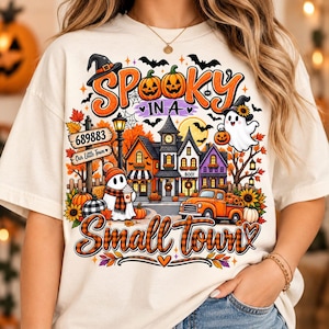 May include: A cream-colored t-shirt featuring a colorful Halloween graphic with the text 'SPOOKY IN A Small town'. The design includes a small town scene with houses, a ghost, an orange truck, pumpkins, sunflowers, and bats under a full moon.
