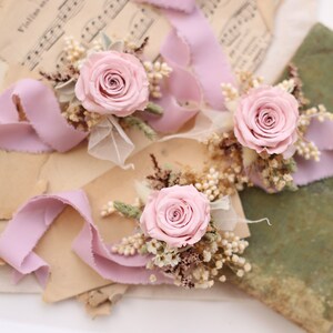 May include: Three floral arrangements featuring light pink roses, dried flowers, and lavender ribbons. The arrangements are set on vintage sheet music and aged paper, creating a romantic and rustic aesthetic.