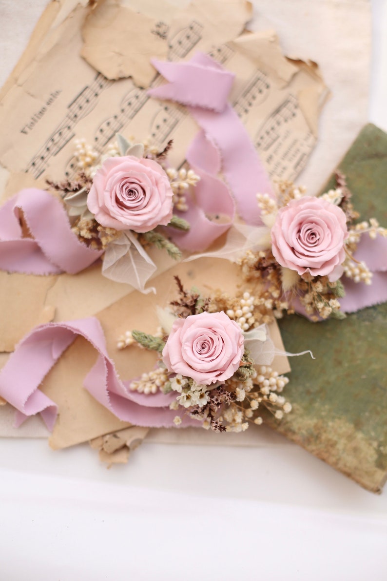 May include: Three floral arrangements featuring light pink roses, dried flowers, and lavender ribbons. The arrangements are set on vintage sheet music and aged paper, creating a romantic and rustic aesthetic.