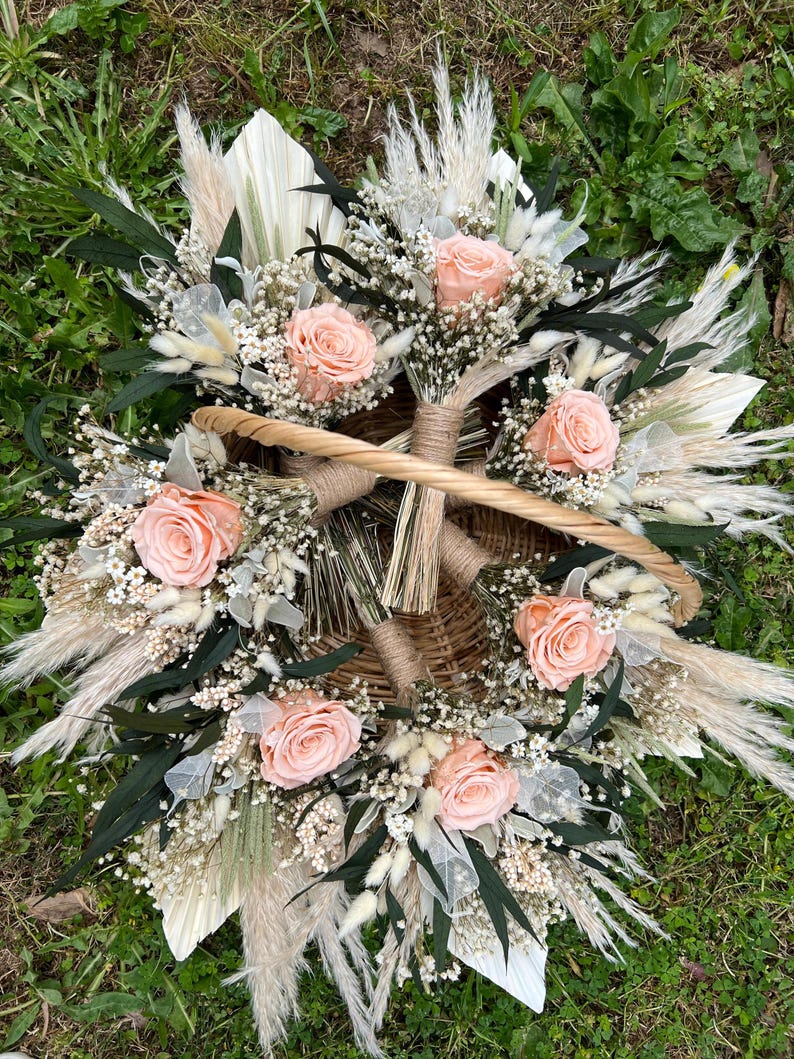 K&ouml;nnte beinhalten: Ein Weidenkorb gef&uuml;llt mit pfirsichfarbenen Rosen, wei&szlig;em Schleierkraut und getrocknetem Pampasgras. Das Arrangement umfasst dunkelgr&uuml;ne Bl&auml;tter und ist auf gr&uuml;nem Gras platziert. Der Korb hat einen geflochtenen Griff.