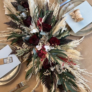 May include: A long, flowing centerpiece made of dried flowers and pampas grass. The centerpiece features burgundy roses, white hydrangeas, and green eucalyptus leaves. The pampas grass is a light beige color. The centerpiece is arranged on a table with two place settings.