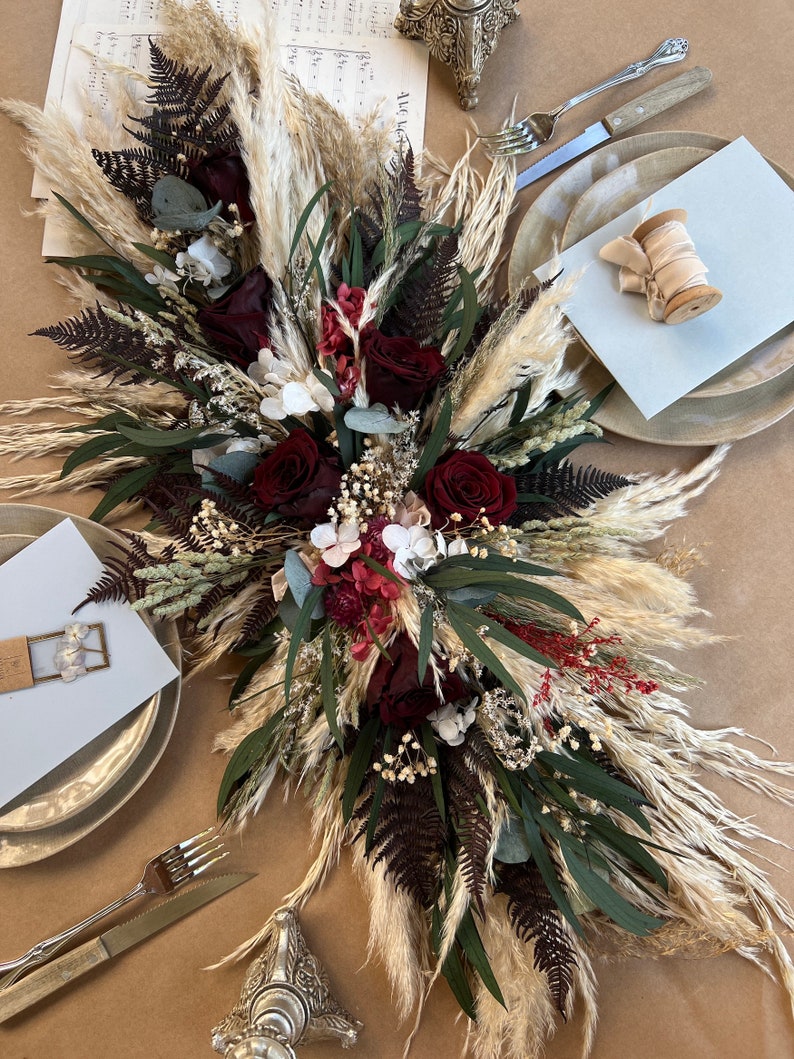 May include: A long, flowing centerpiece made of dried flowers and pampas grass. The centerpiece features burgundy roses, white hydrangeas, and green eucalyptus leaves. The pampas grass is a light beige color. The centerpiece is arranged on a table with two place settings.