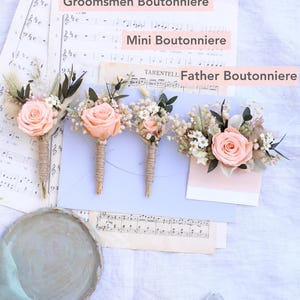 K&ouml;nnte beinhalten: Vier Boutonnieres mit pfirsichfarbenen Rosen und Gr&uuml;nzeug, angeordnet auf Notenbl&auml;ttern. Die Boutonnieres sind mit "Groom Boutonniere", "Groomsmen Boutonniere", "Mini Boutonniere" und "Father Boutonniere" beschriftet. Jede Boutonniere ist mit Bindfaden umwickelt.