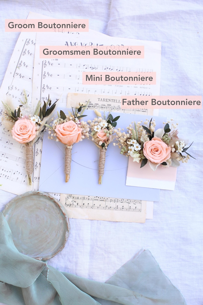 K&ouml;nnte beinhalten: Vier Boutonnieres mit pfirsichfarbenen Rosen und Gr&uuml;nzeug, angeordnet auf Notenbl&auml;ttern. Die Boutonnieres sind mit "Groom Boutonniere", "Groomsmen Boutonniere", "Mini Boutonniere" und "Father Boutonniere" beschriftet. Jede Boutonniere ist mit Bindfaden umwickelt.