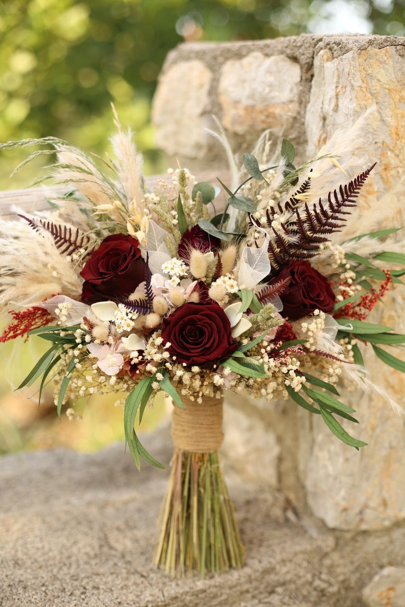 May include: A burgundy and cream dried flower bouquet with pampas grass, baby's breath, and red roses. The bouquet is tied with twine and has a rustic, bohemian style.
